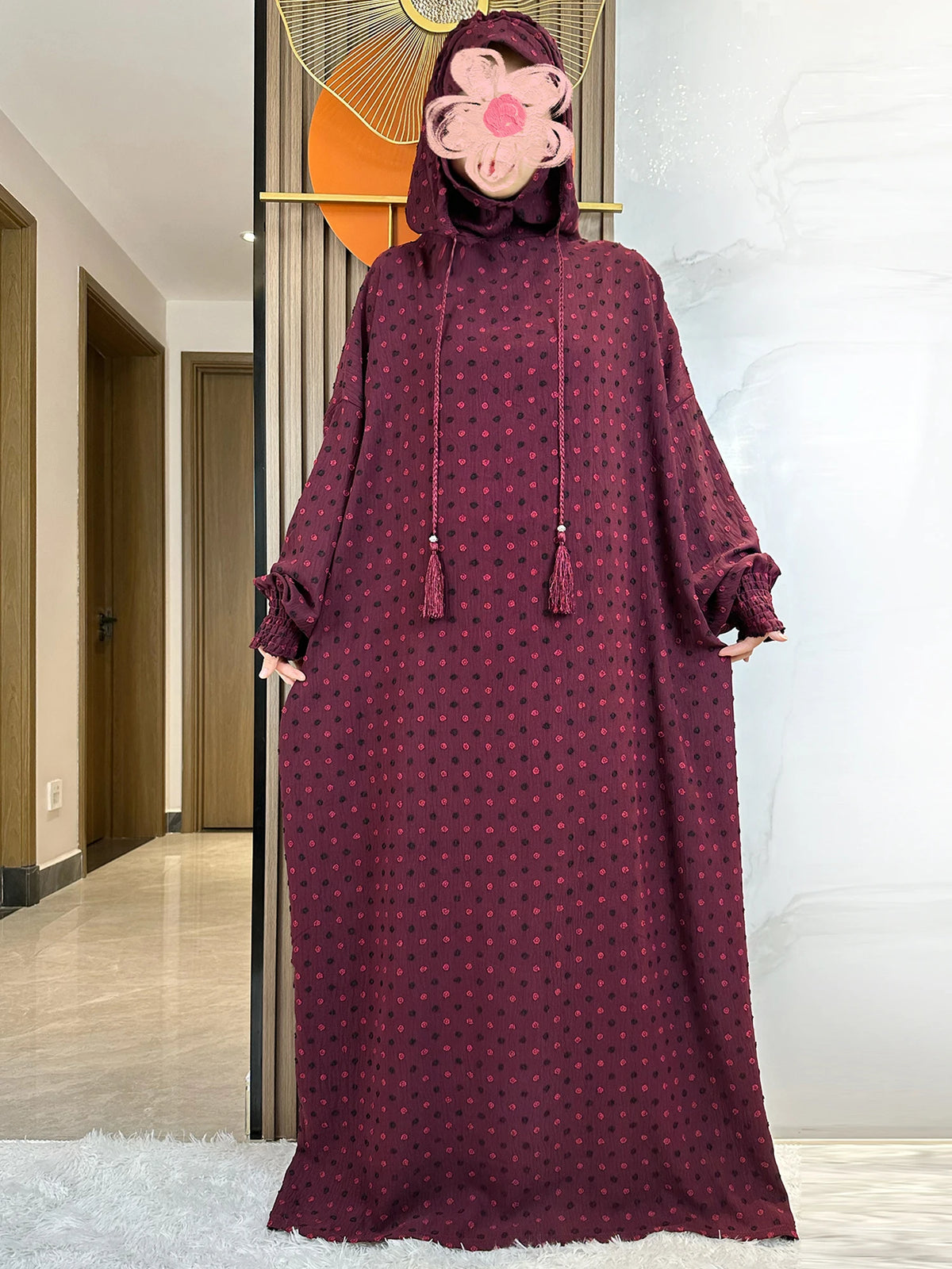 Abaya de algodón color granate con detalle floral rosa, exhibida contra una pared blanca.