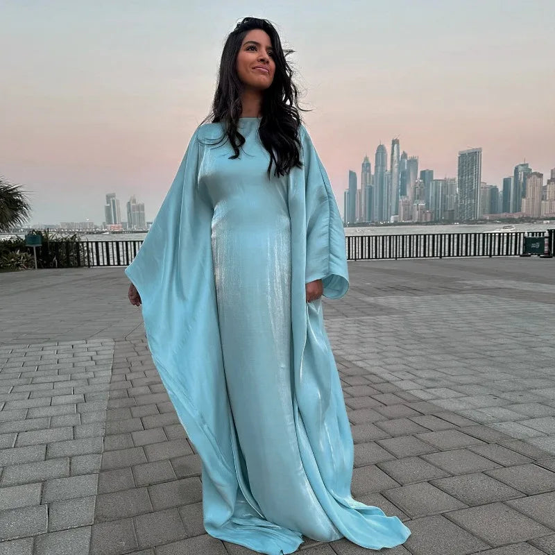 Eine Frau in einem eleganten, fließenden, hellblauen Langarm-Abaya steht draußen vor einer Stadtansicht.
