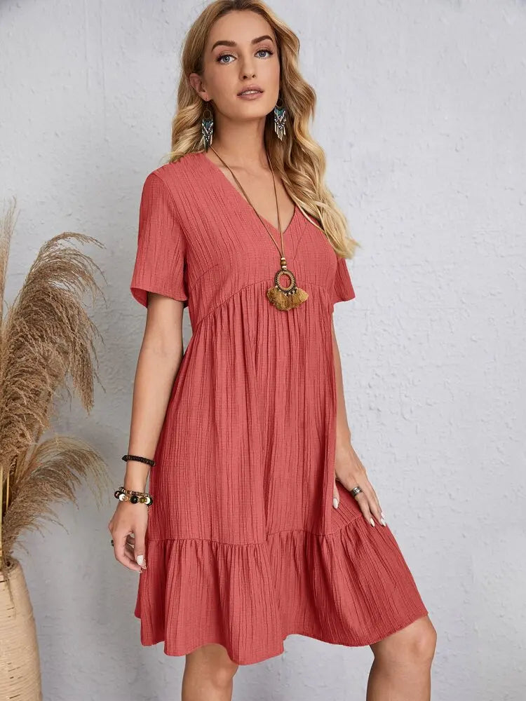 Eine Frau modelliert ein korallenrosa, einfaches V-Ausschnitt, A-Linien-Minikleid mit goldenen Ohrringen und Armreifen.