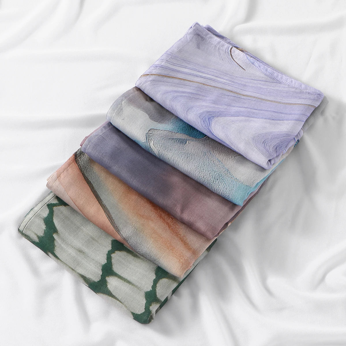 A stack of five watercolor print modal scarves in shades of purple, blue, gray, and peach laid on white fabric.