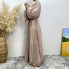 Una mujer vistiendo un cárdigan abaya de cuentas beige, de pie contra una pared blanca con hierbas secas y una pintura abstra