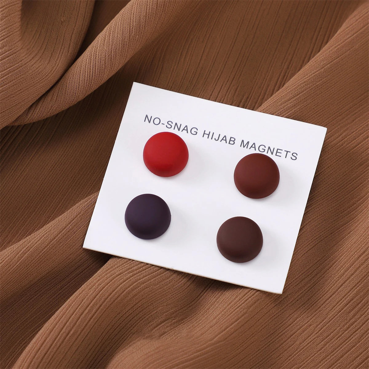 Weiße Karte mit fünf runden magnetischen Hijab-Pins in Rot-, Braun- und Schwarztönen auf drapiertem Stoff.