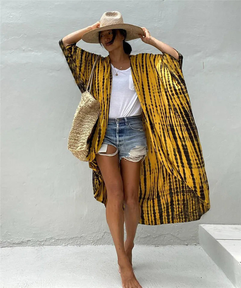 Eine Frau trägt einen gelb-schwarz gestreiften Kimono-Überwurf über einem weißen Oberteil und Jeansshorts und hält einen Stro