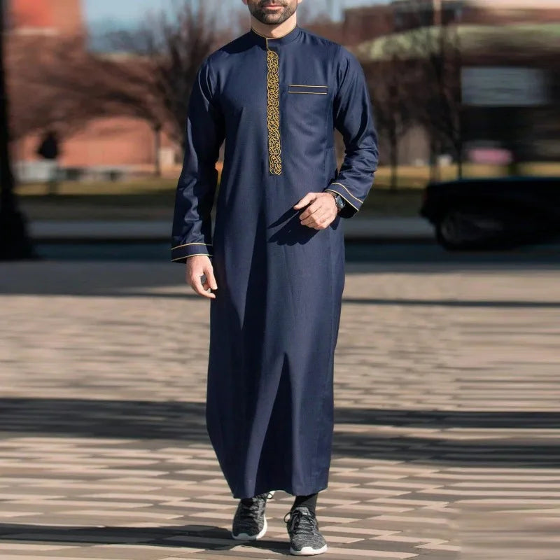 Un hombre vistiendo una jubba thobe bordada azul marino, bata casual, de pie al aire libre.