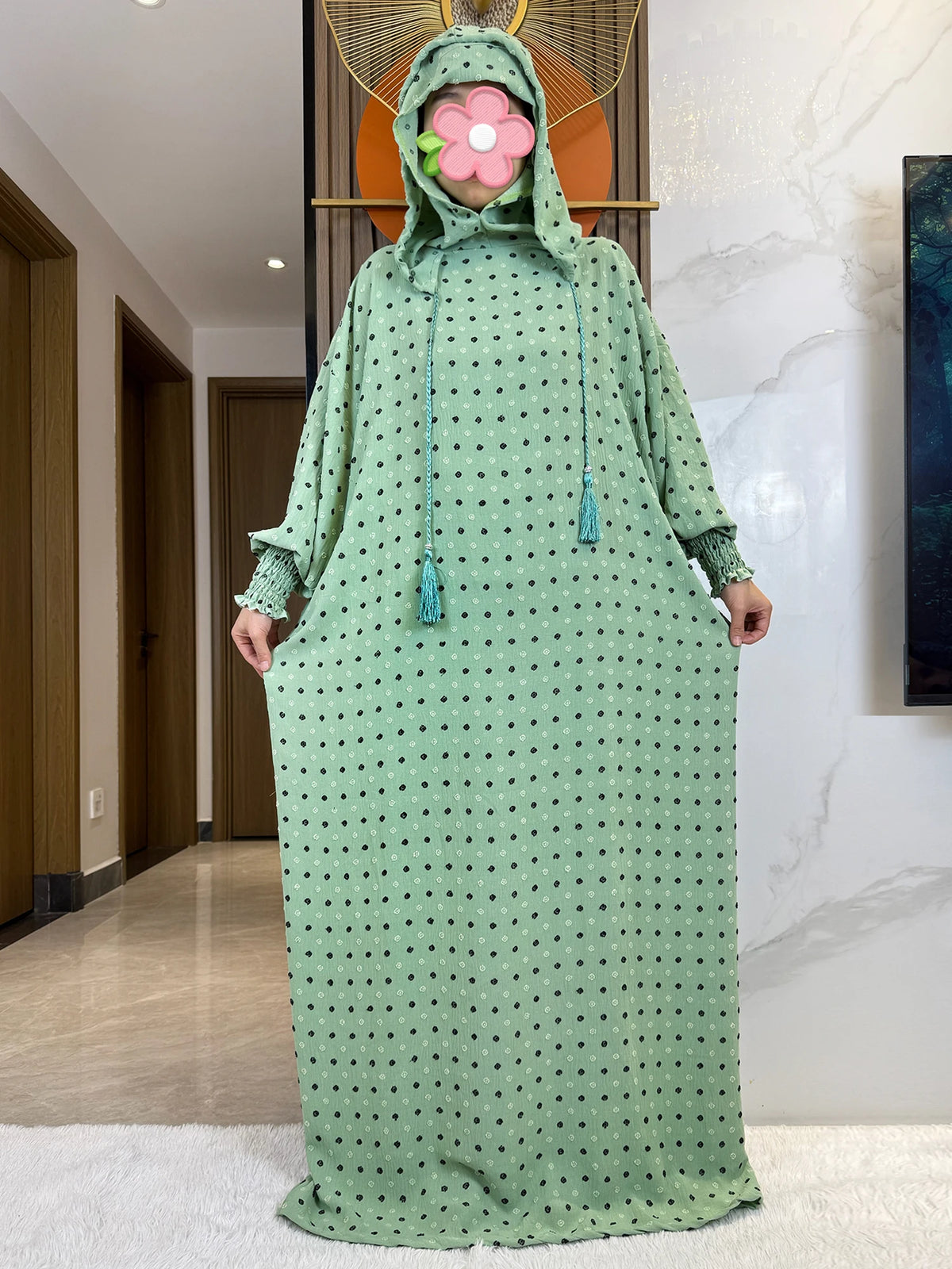 Una mujer vistiendo una abaya de algodón verde oliva larga con capucha y detalle floral, de pie en un pasillo.