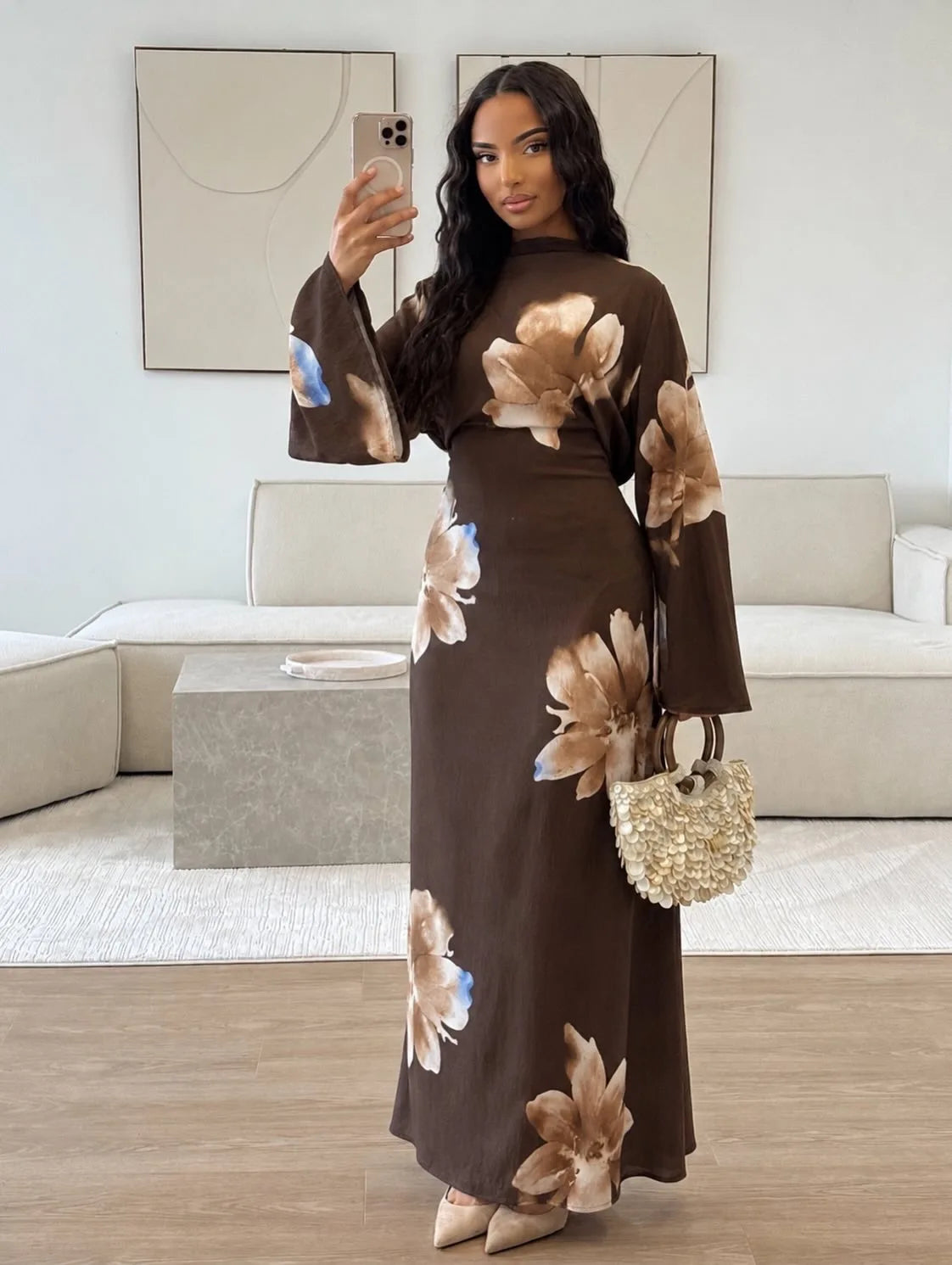 Eine Frau modelliert ein langes, dunkelbraunes Abaya-Kleid mit großen Blumenmustern in Beige und Blau vor hellem Hintergrund.