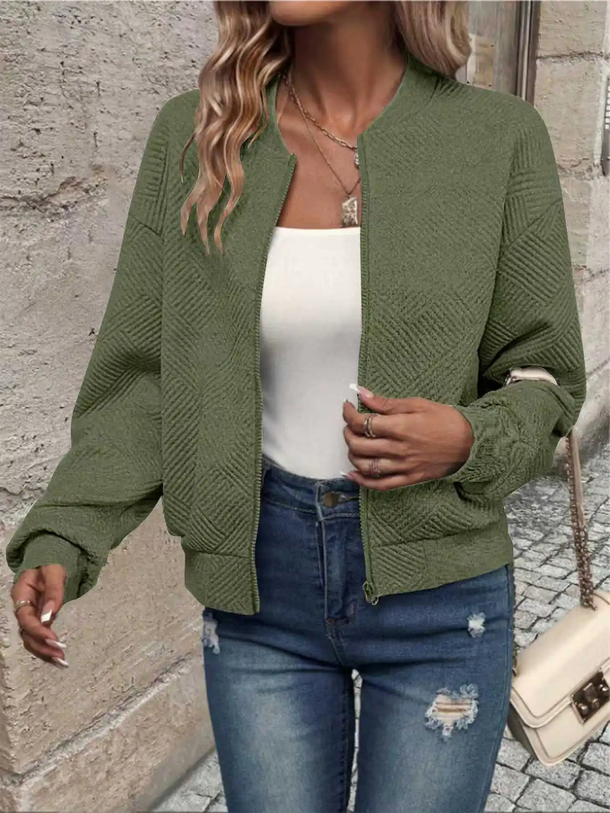 Une femme portant une veste à fermeture éclair tricotée vert olive sur un haut blanc et un jean bleu.