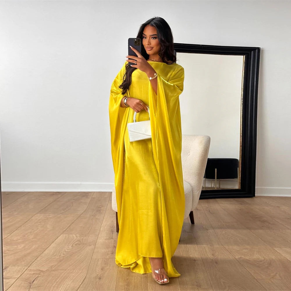 Una mujer vistiendo una abaya de satén amarillo brillante con diseño de ala y sosteniendo un teléfono frente a un gran espejo