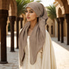 Una mujer con hijab beige y prenda blanca fluida, de pie en un pasillo exterior con palmeras.