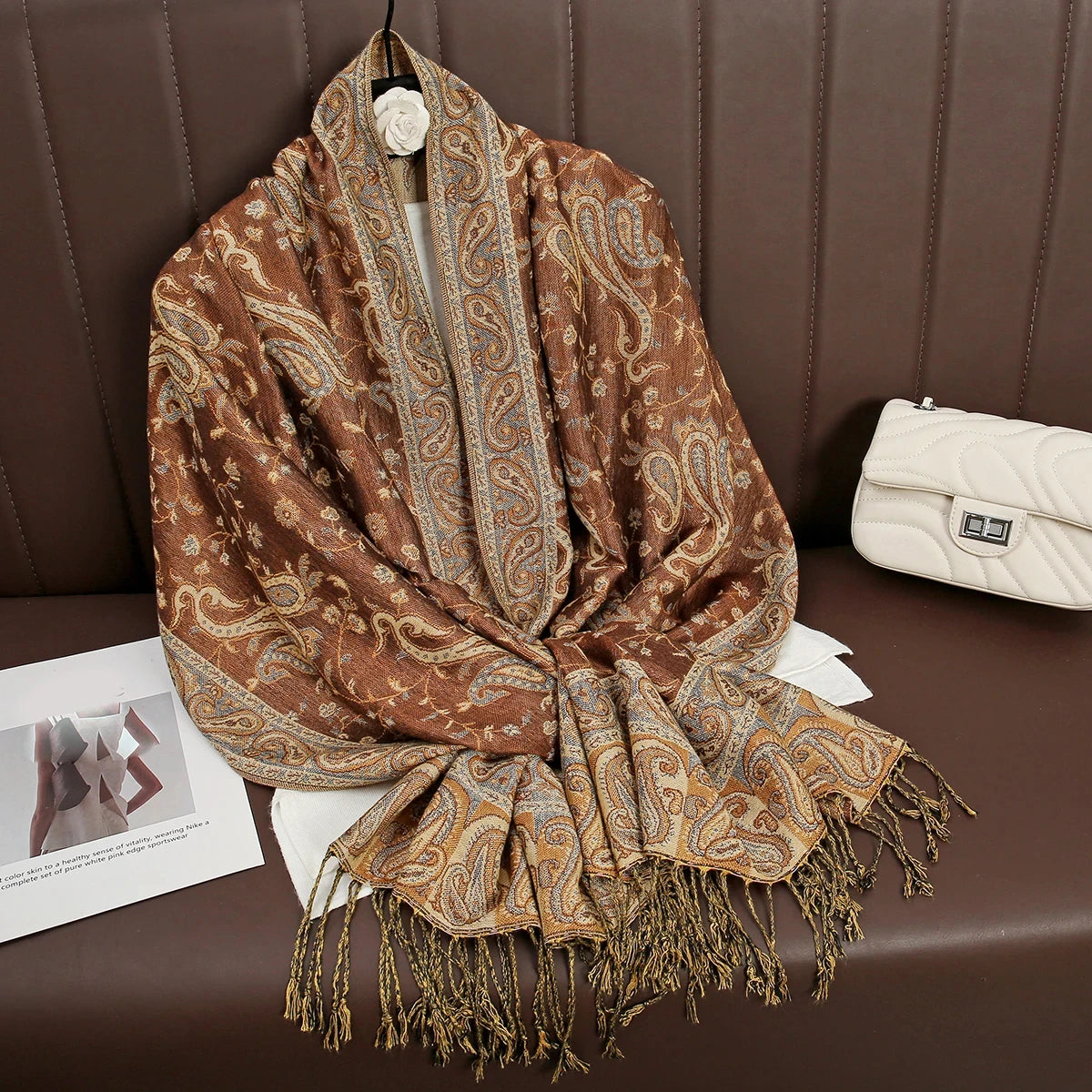 Pañuelo de pashmina de lujo marrón estampado drapeado sobre una superficie de cuero marrón junto a un clutch color crema.