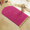 Alfombra de oración magenta con patrón geométrico grabado y borlas doradas sobre un suelo de madera clara.