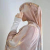 Eine Frau mit einem durchscheinenden rosa Hijab mit goldenen Aquarellakzenten, die über ihre Schulter blickt.