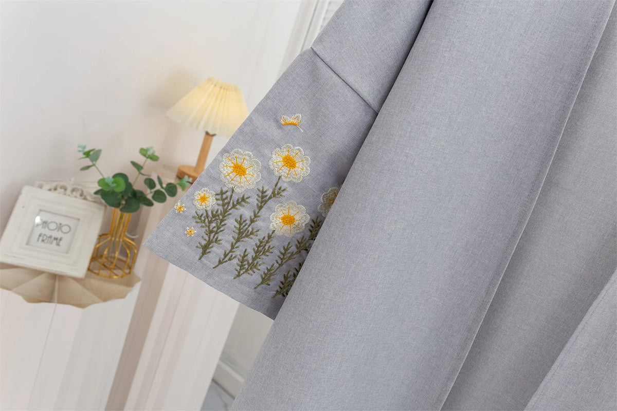 Graues Stoffstück mit gelber Blumenstickerei neben einer kleinen goldenen Vase und einer Lampe.