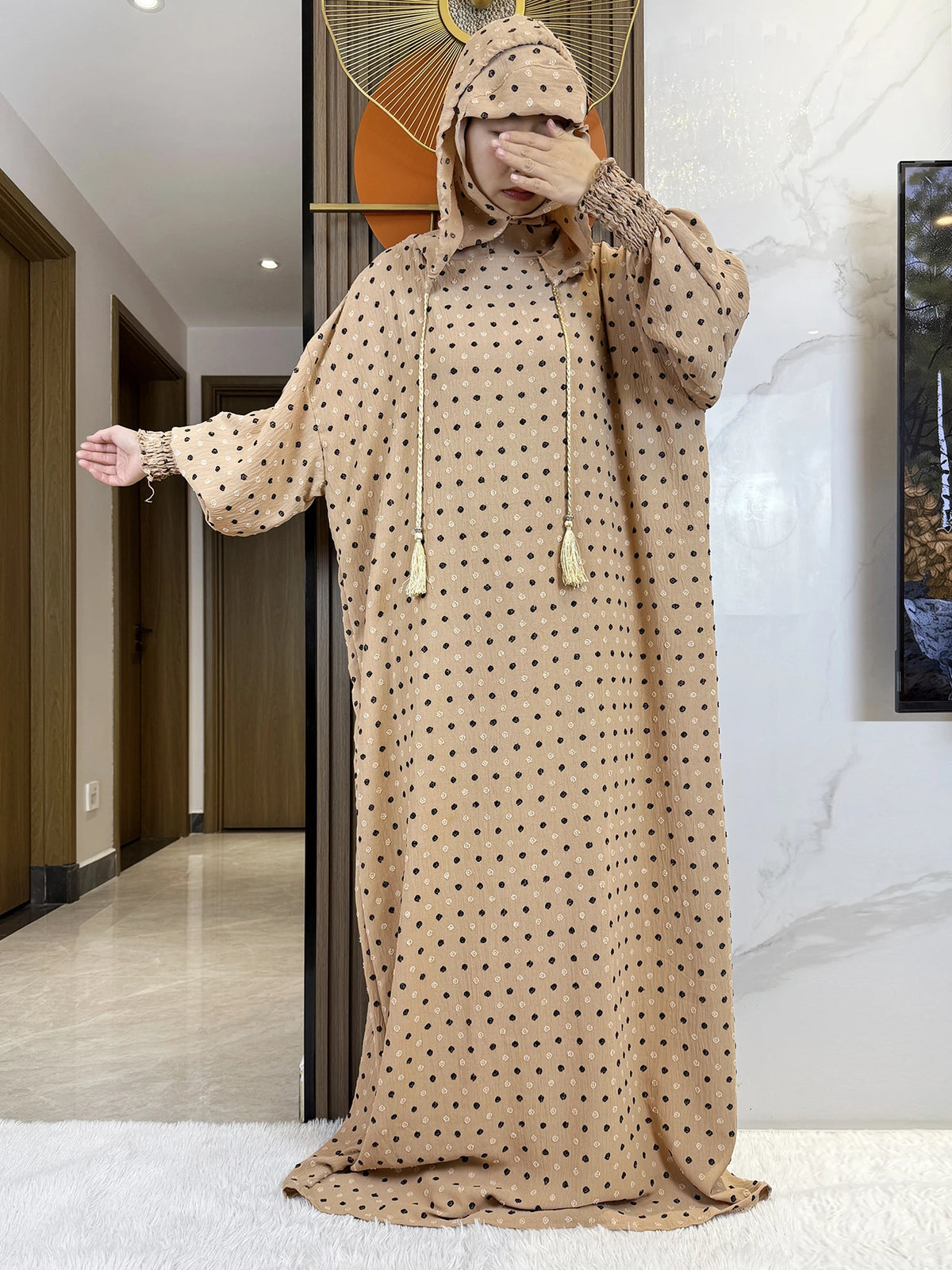 Una mujer vistiendo una abaya de algodón beige con puntos, capucha y velo de oración, está de pie en un pasillo.