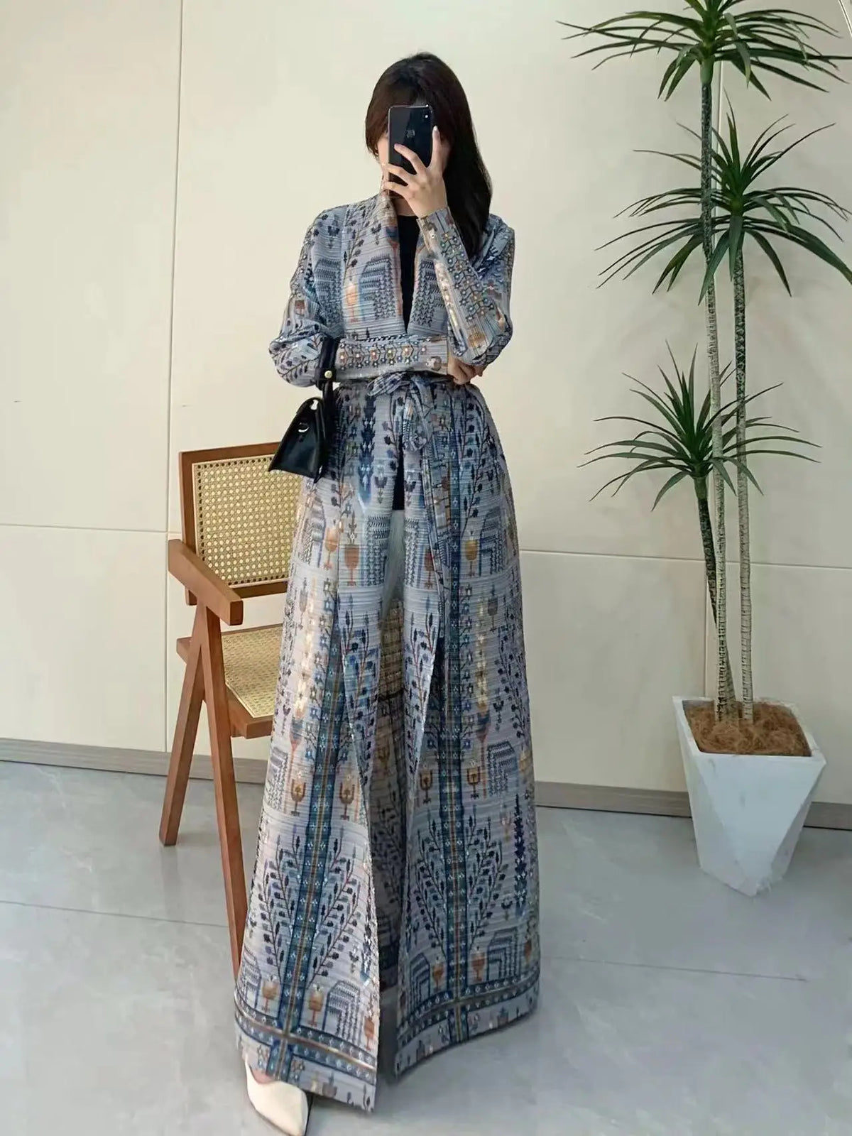 Une femme modélise un trench-coat à manches longues plissé bleu et blanc avec des accessoires noirs dans un intérieur lumineu