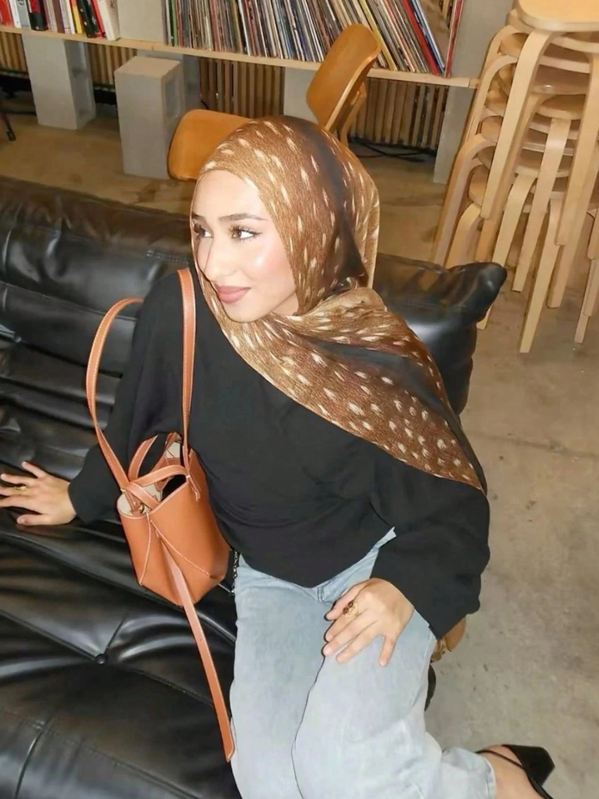 Eine Frau mit braunem Aquarell-Hirschmuster-Hijab-Schal und schwarzem Oberteil sitzt auf einem Ledersofa.