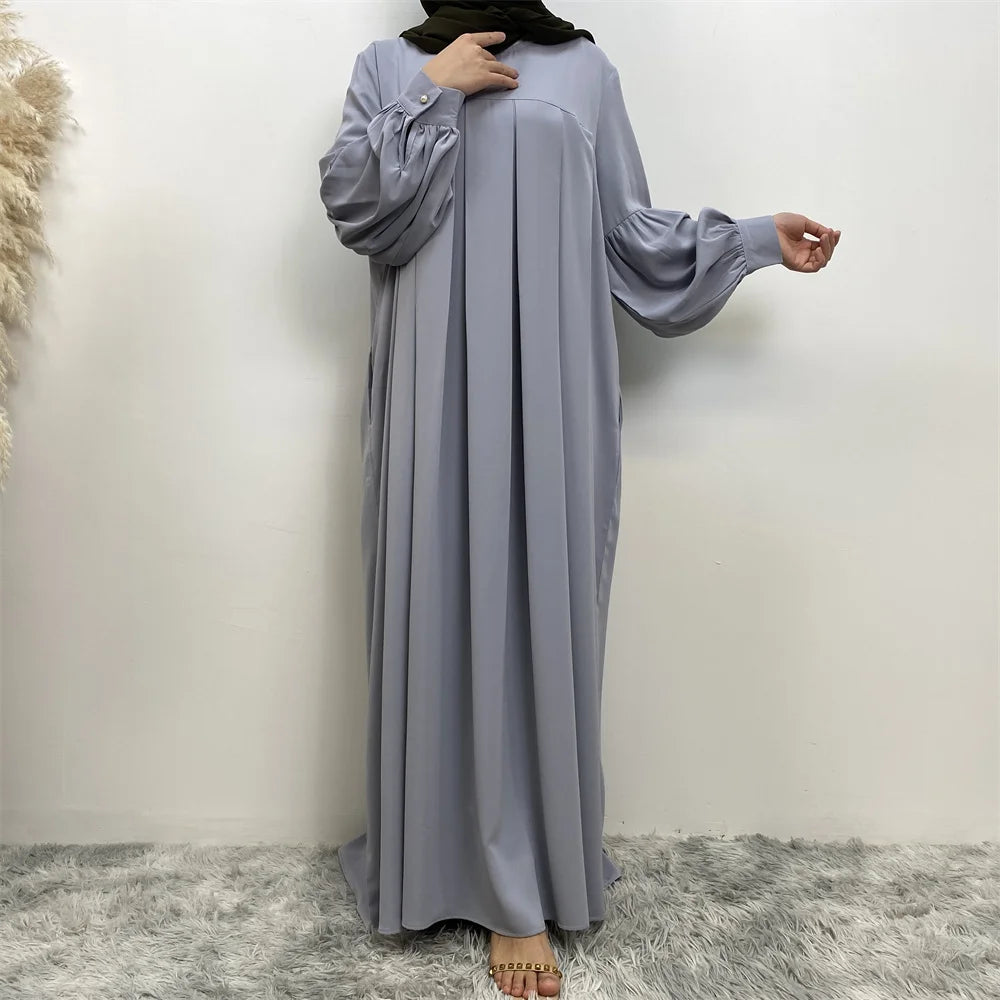 Eine Frau in einem langen, fließenden, hellgrauen islamischen Kleid mit voluminösen Ärmeln und einem schwarzen Hijab.