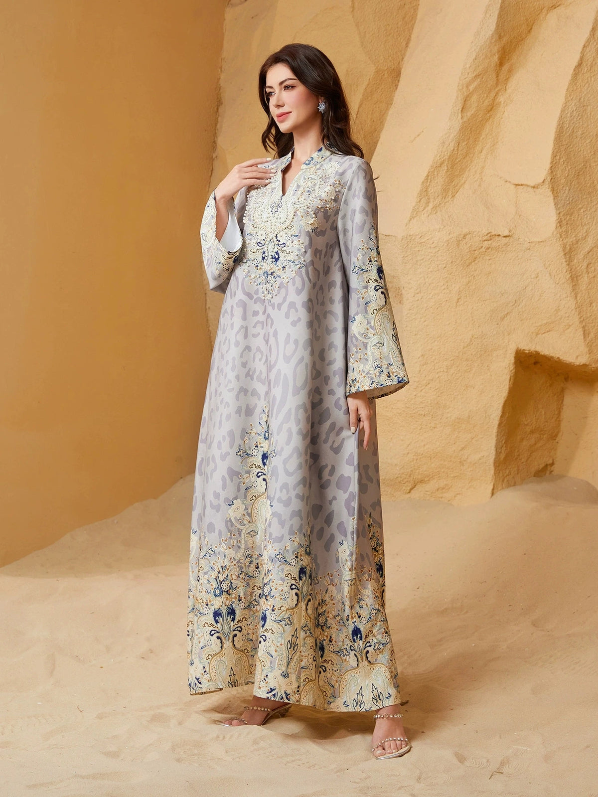 Una mujer vistiendo una abaya larga gris claro con sutil estampado de leopardo y adornos florales azules, de pie en un paisaj