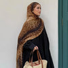 Une femme portant un hijab bohème à imprimé léopard marron et une robe noire, debout à côté d'une porte verte.