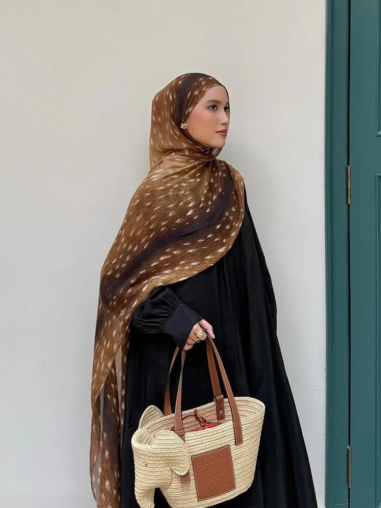Une femme portant un hijab bohème à imprimé léopard marron et une robe noire, debout à côté d'une porte verte.