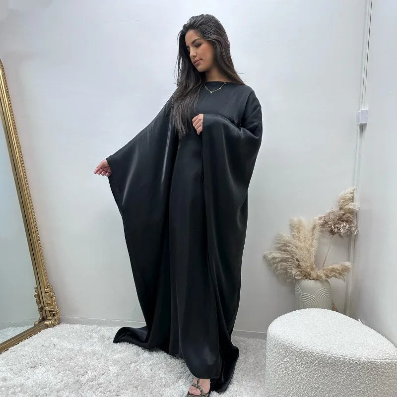 Eine Frau in einer eleganten schwarzen Langarm-Abaya steht vor einer weißen Wand.