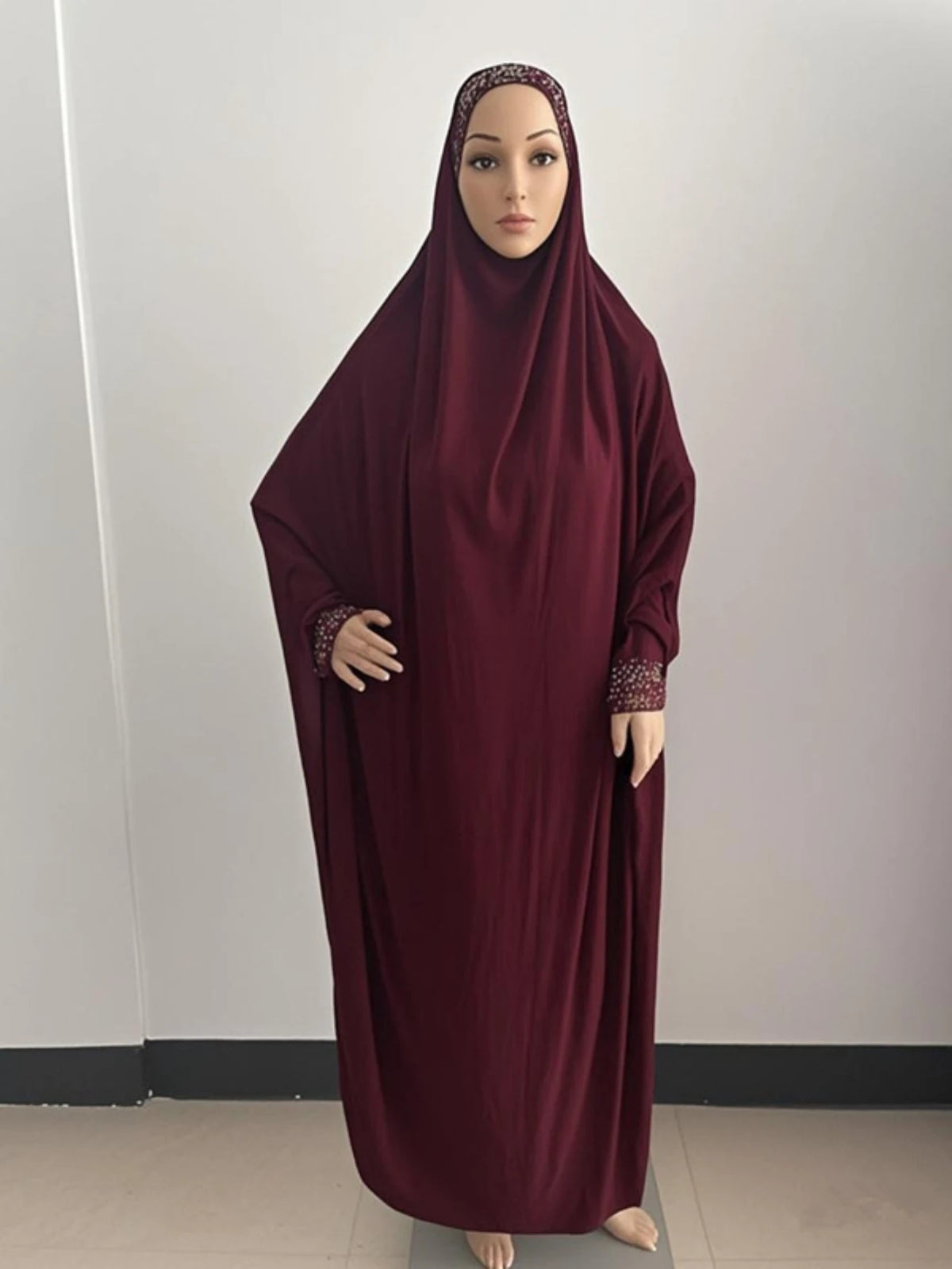 Une femme portant une caftan abaya fluide bordeaux avec un hijab, debout sur un fond clair.