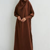 Eine Frau in einem braunen, bescheidenen Abaya mit gerafften Ärmeln, vor hellem Hintergrund.