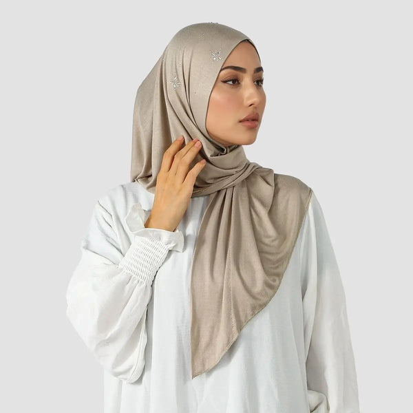 A woman wearing a beige triangle jersey hijab scarf and white top, looking to the side.