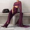 A model wears a sheer, long-sleeved plum mesh poncho cape over a dark purple jumpsuit in an indoor setting.