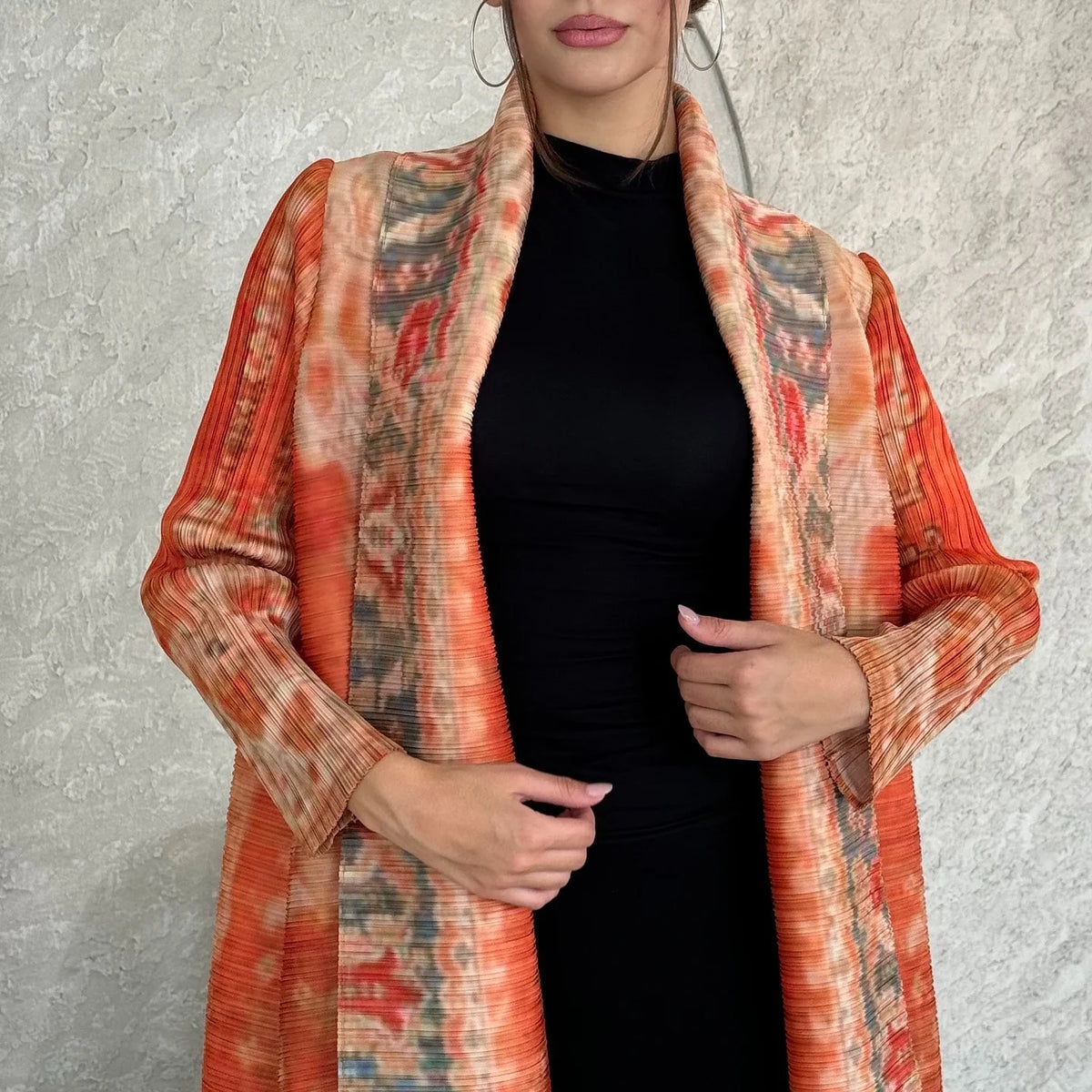 Eine Frau trägt einen orange-beige gemusterten, fließenden, plissierten Abaya-Cardigan über einem schwarzen Kleid.