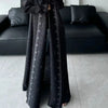 Une femme portant un long trench-coat plissé noir à motifs, debout à l'intérieur.