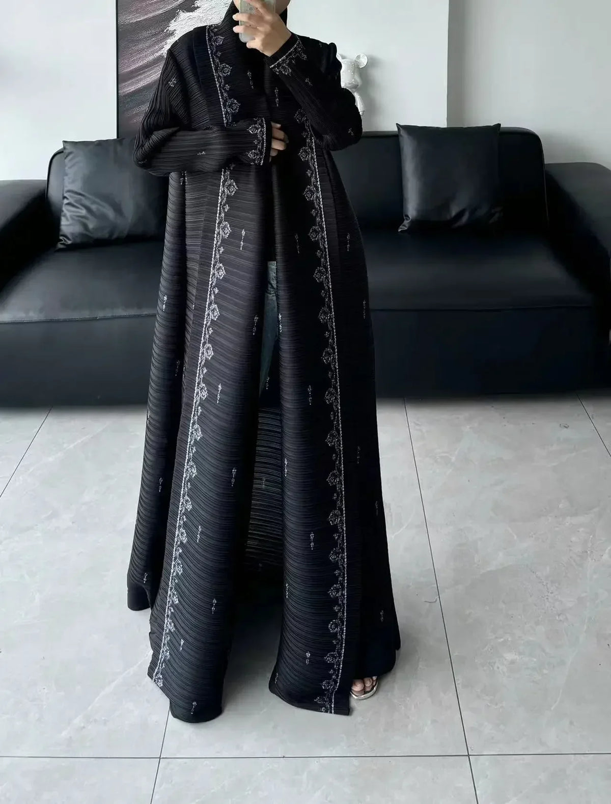 Une femme portant un long trench-coat plissé noir à motifs, debout à l'intérieur.