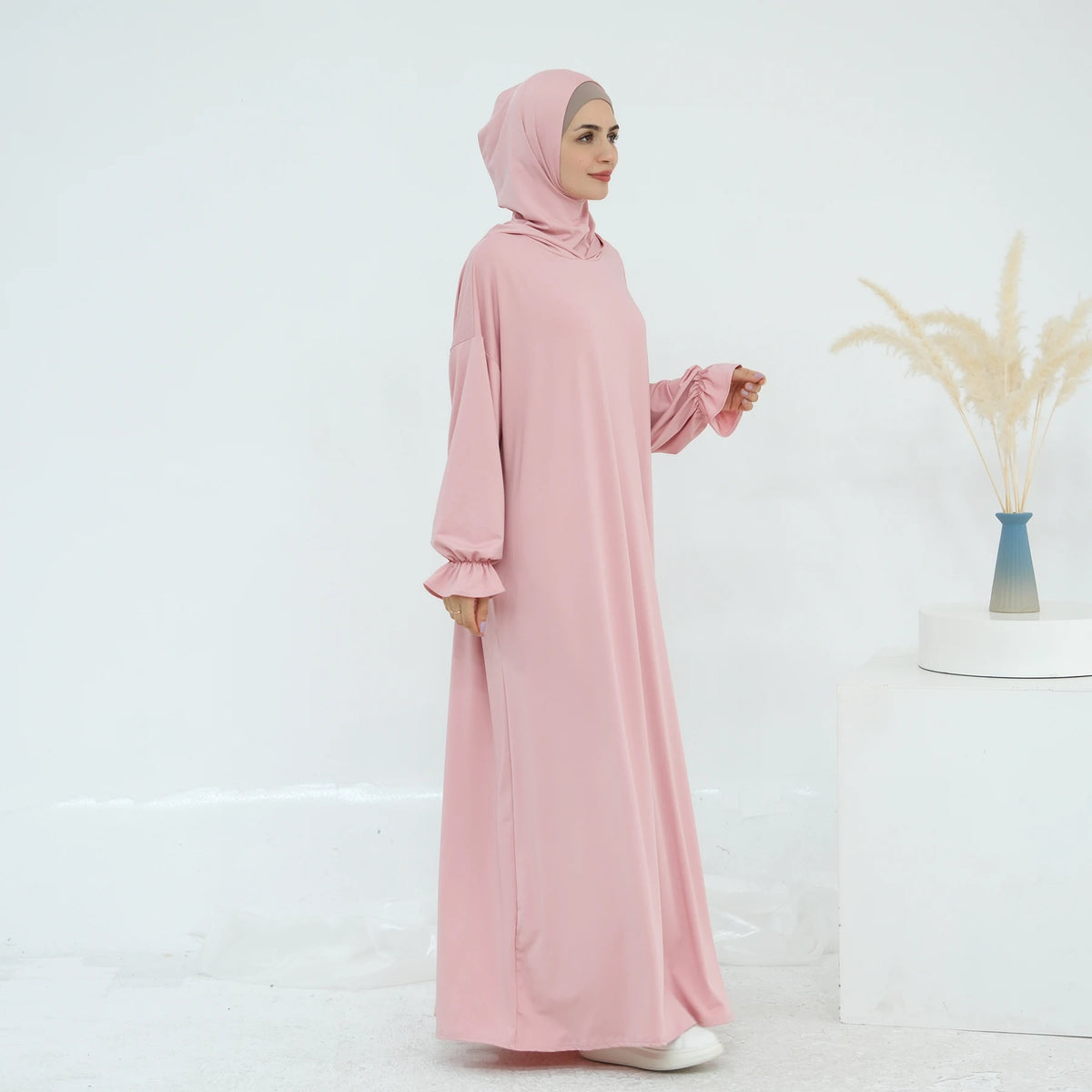 Una mujer vistiendo un vestido largo y suelto de color rosa para la oración, de pie contra un fondo blanco.