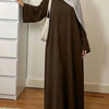 Une femme portant une élégante abaya brune ample et un hijab gris clair, à côté d'une armoire blanche.