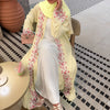 Une femme portant une abaya jaune pâle avec des détails en dentelle florale et un foulard assorti, assise à l'intérieur.