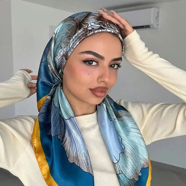 A woman models a blue and gold printed satin silk hijab scarf against a light background.