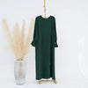 Vestido abaya verde oscuro y holgado colgado en percha de madera sobre fondo blanco.