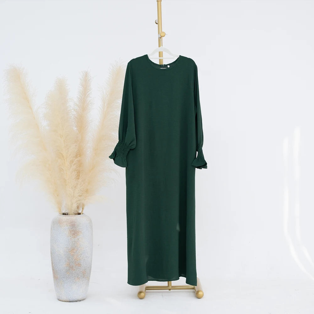 Vestido abaya verde oscuro y holgado colgado en percha de madera sobre fondo blanco.