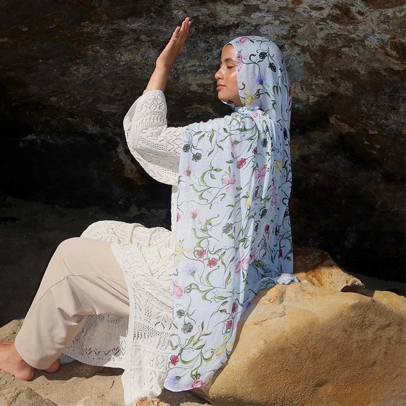 Une femme portant un haut tricoté blanc et un pantalon beige clair, drapée d'un hijab floral aquarelle.