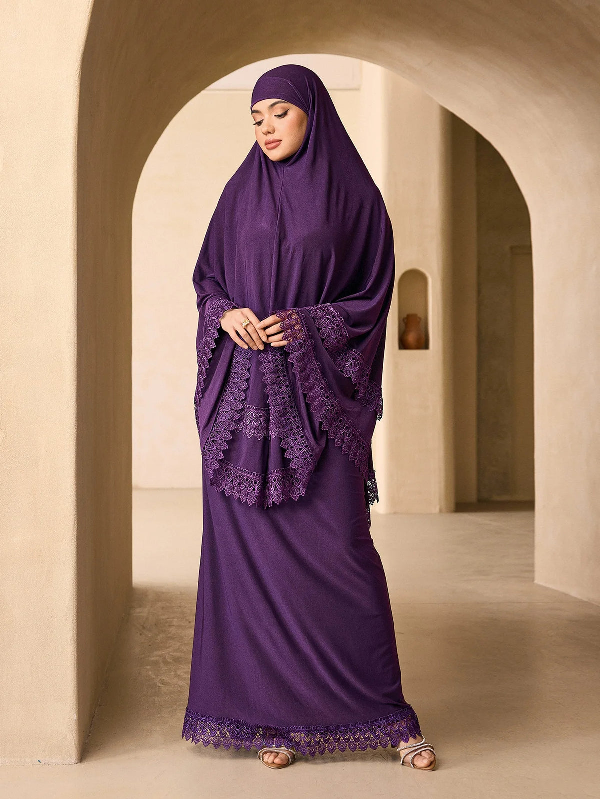 Eine Frau in einem eleganten, mehrlagigen Spitzen-Khimar-Abaya-Set in tiefem Violett steht in einem hellen, gewölbten Flur.