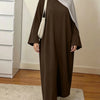 Une femme portant une abaya maxi ample marron et un hijab gris se tient dans une pièce bien éclairée.