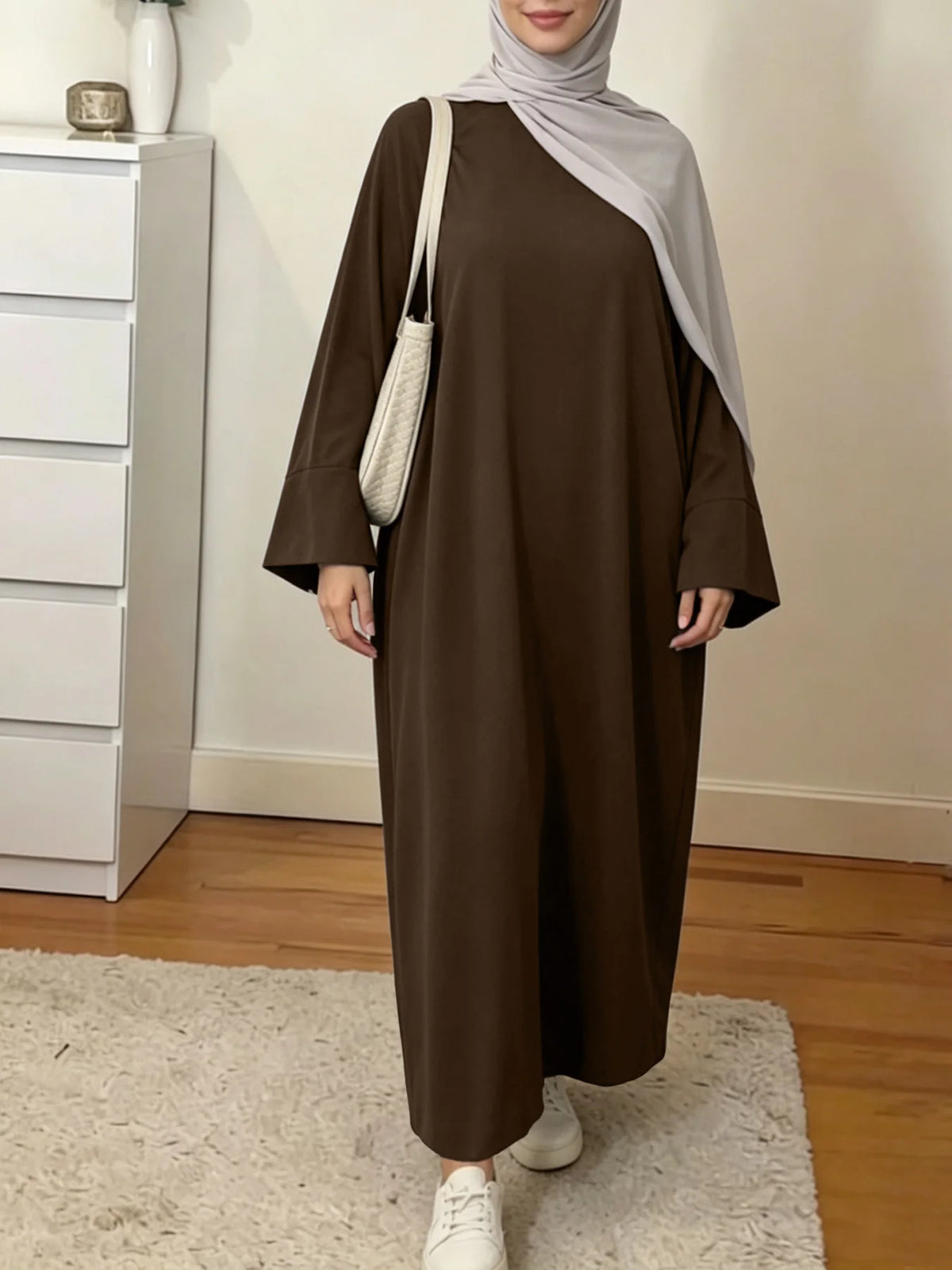 Une femme portant une abaya maxi ample marron et un hijab gris se tient dans une pièce bien éclairée.