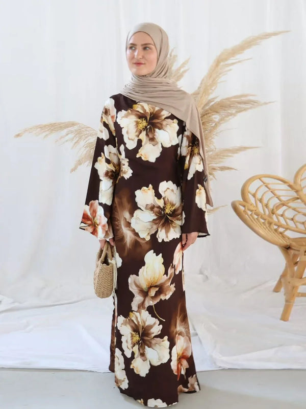 A woman wearing a brown and cream floral print abaya with a hijab stands in front of a white background.