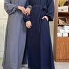 Eine Frau in einem dunkelblauen marokkanischen Kaftan-Abaya mit grauem Hijab vor Holzregalen.