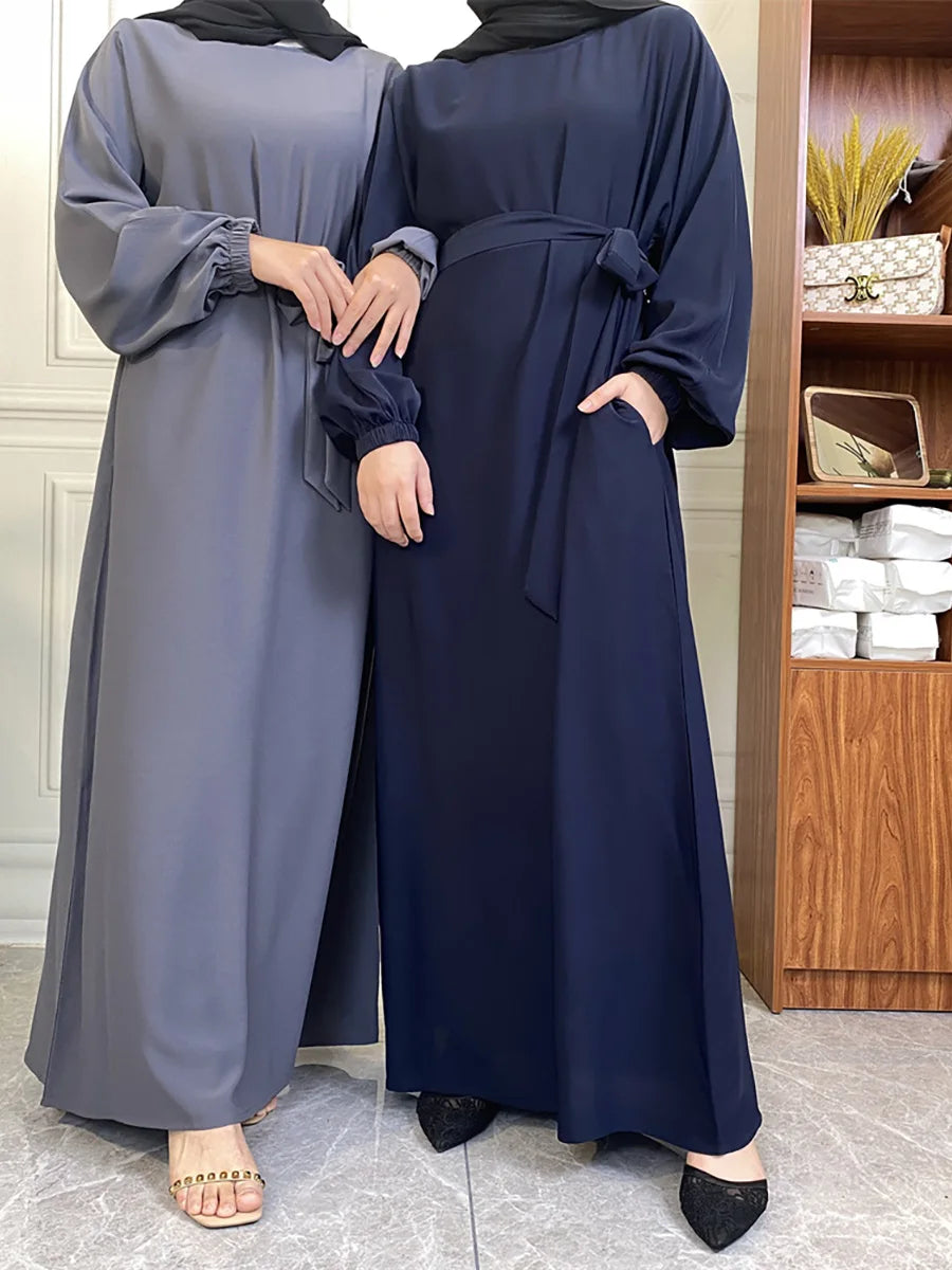 Eine Frau in einem dunkelblauen marokkanischen Kaftan-Abaya mit grauem Hijab vor Holzregalen.