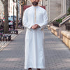 A man wearing a white Moroccan ethnic thobe with a stand collar standing on a sidewalk.