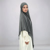 A woman wearing a gray square hijab and white clothing is shown in a full-length portrait.