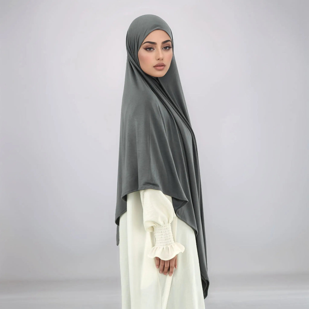 A woman wearing a gray square hijab and white clothing is shown in a full-length portrait.