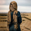 Una mujer con hijab tie-dye en degradado beige y chaqueta de mezclilla, de pie en un campo seco bajo un cielo nublado.