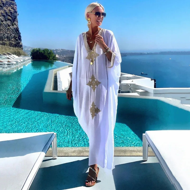 Eine Frau in einem weißen Kaftan mit goldenen Stickereien steht aminfty Pool mit Blick auf den Ozean.